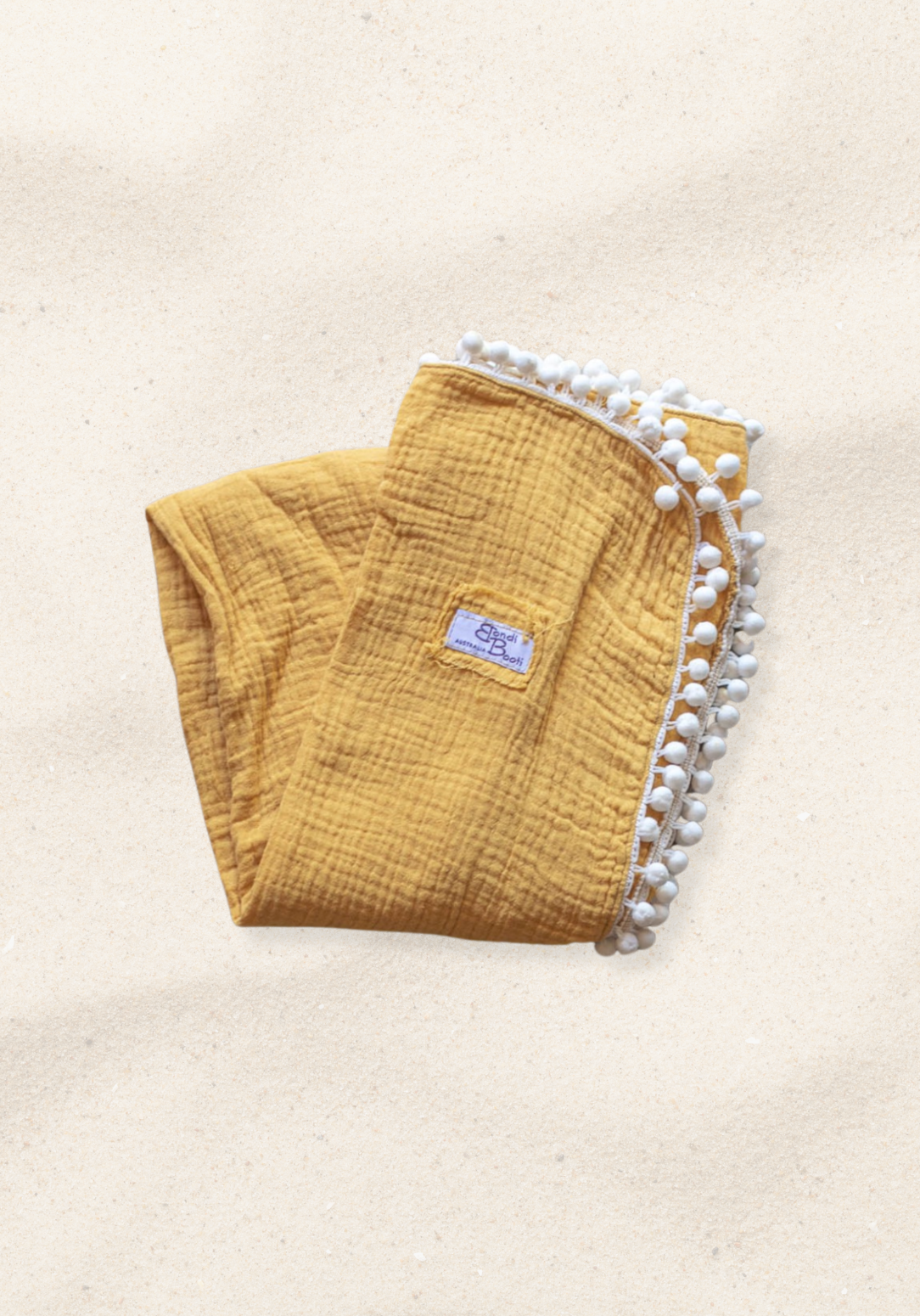 Mustard yellow organic cotton baby blanket with white pom-pom trim, folded neatly with a Bondi Booti label, placed on a neutral surface.