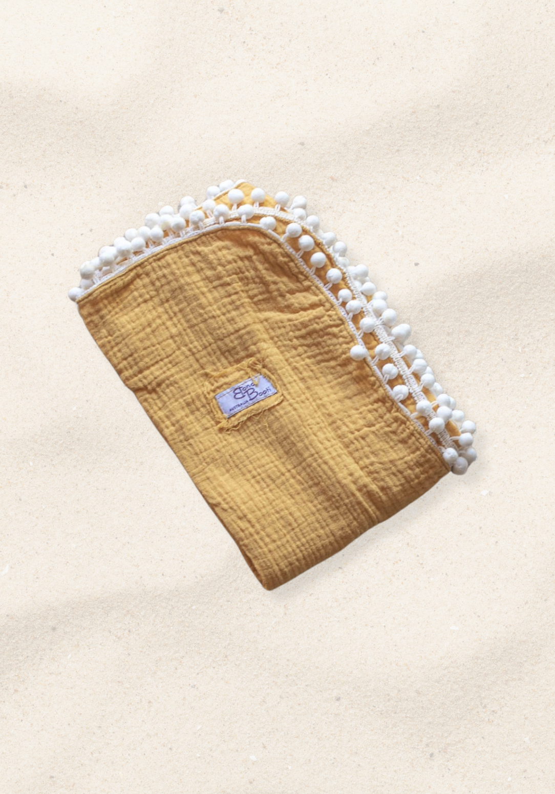 Mustard yellow organic cotton baby blanket with white pom-pom trim, folded neatly with a Bondi Booti label, placed on a neutral surface.