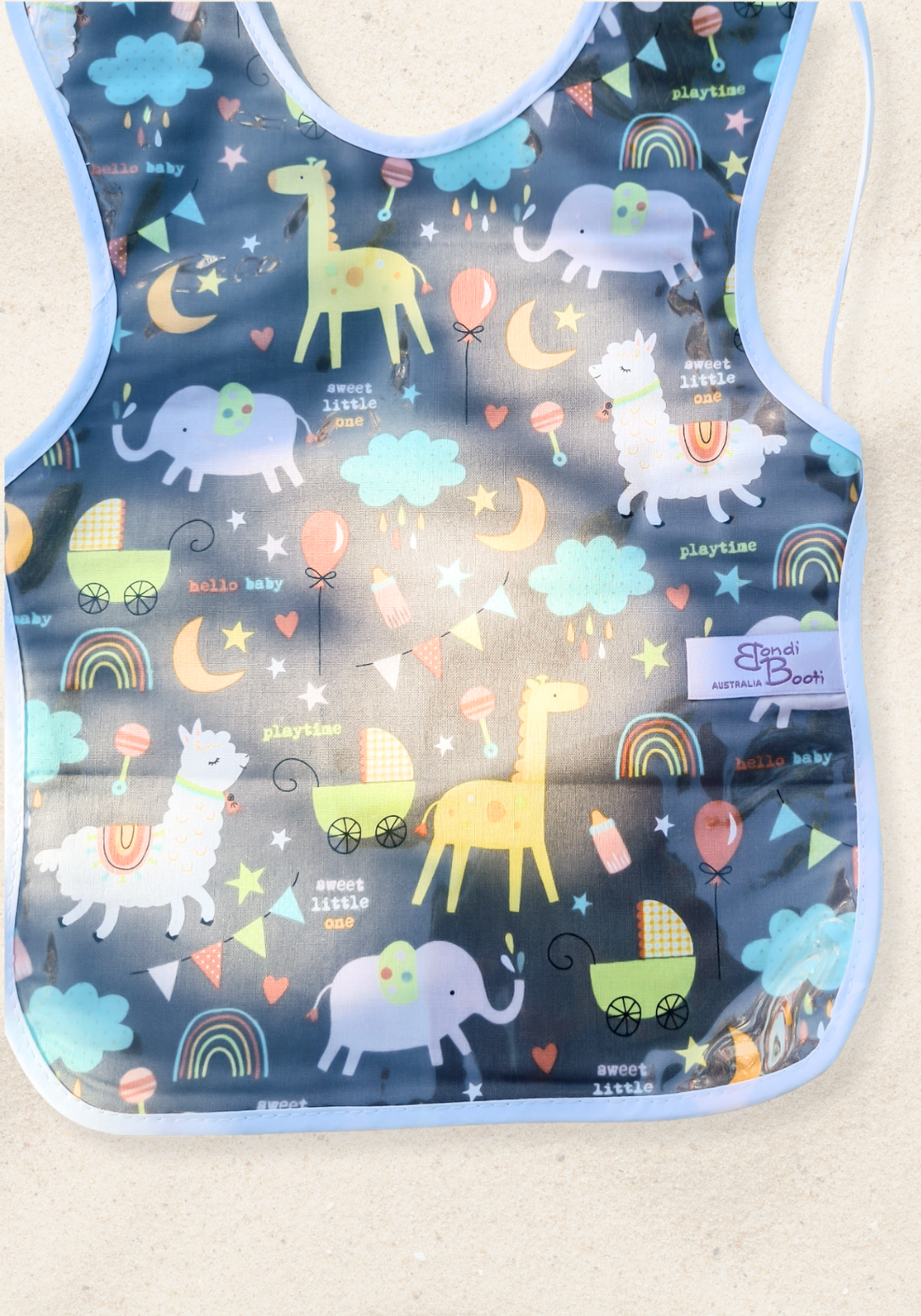 Laminated toddler feeding bib with a whimsical nighttime animal design, featuring giraffes, elephants, rainbows, and clouds on a dark grey background, with white trim and tie closure, laid flat on a sand background.
