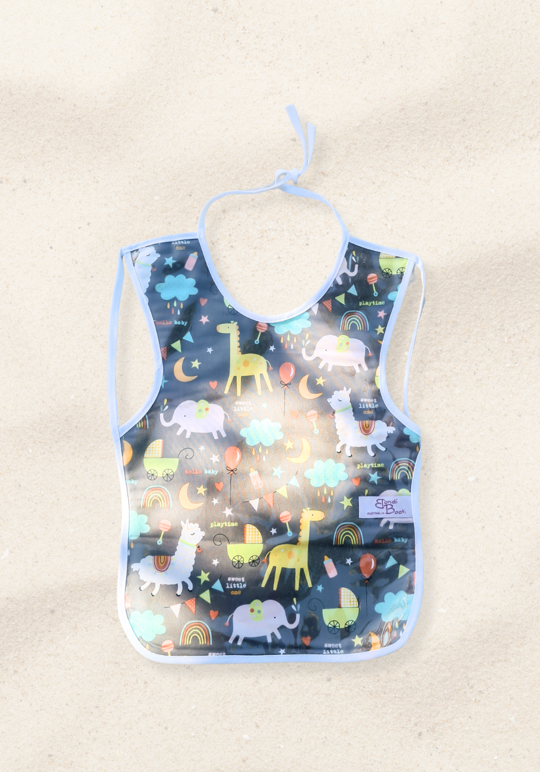 Laminated toddler feeding bib with a whimsical nighttime animal design, featuring giraffes, elephants, rainbows, and clouds on a dark grey background, with white trim and tie closure, laid flat on a sand background.