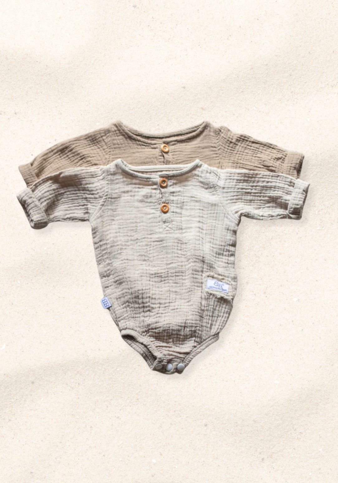 Beige long sleeve baby romper made from organic cotton with wooden buttons and snap closure, laid flat on a sand-colored background