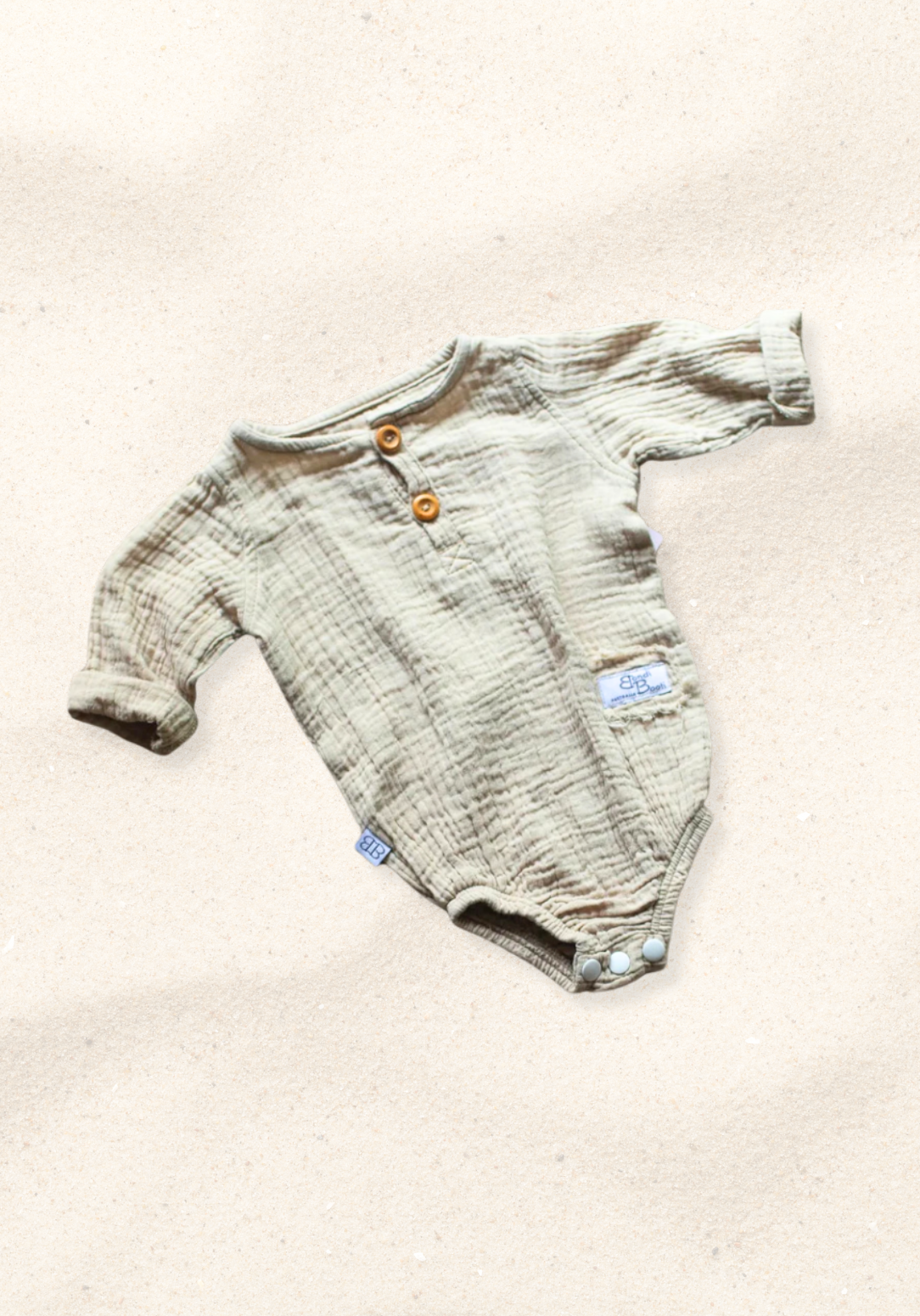 Beige long sleeve baby romper made from organic cotton with wooden buttons and snap closure, laid flat on a sand-colored background