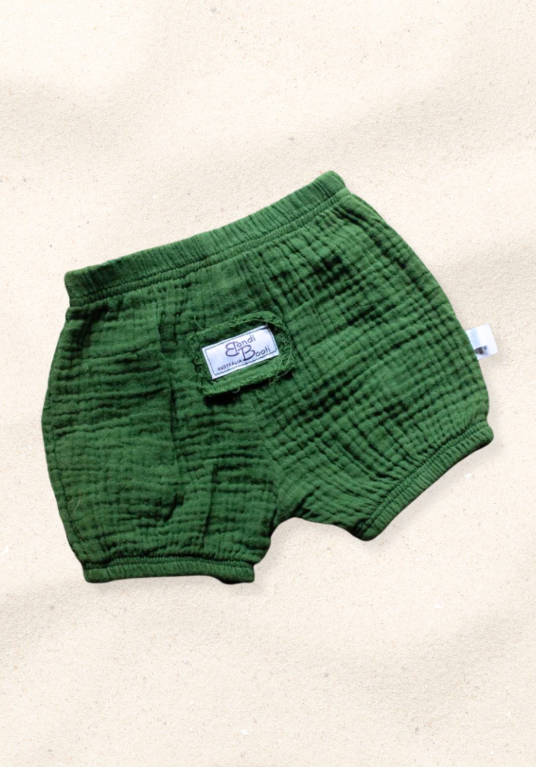 Deep green organic cotton baby bloomers with elastic waistband and leg openings, laid flat on a sand-colored background