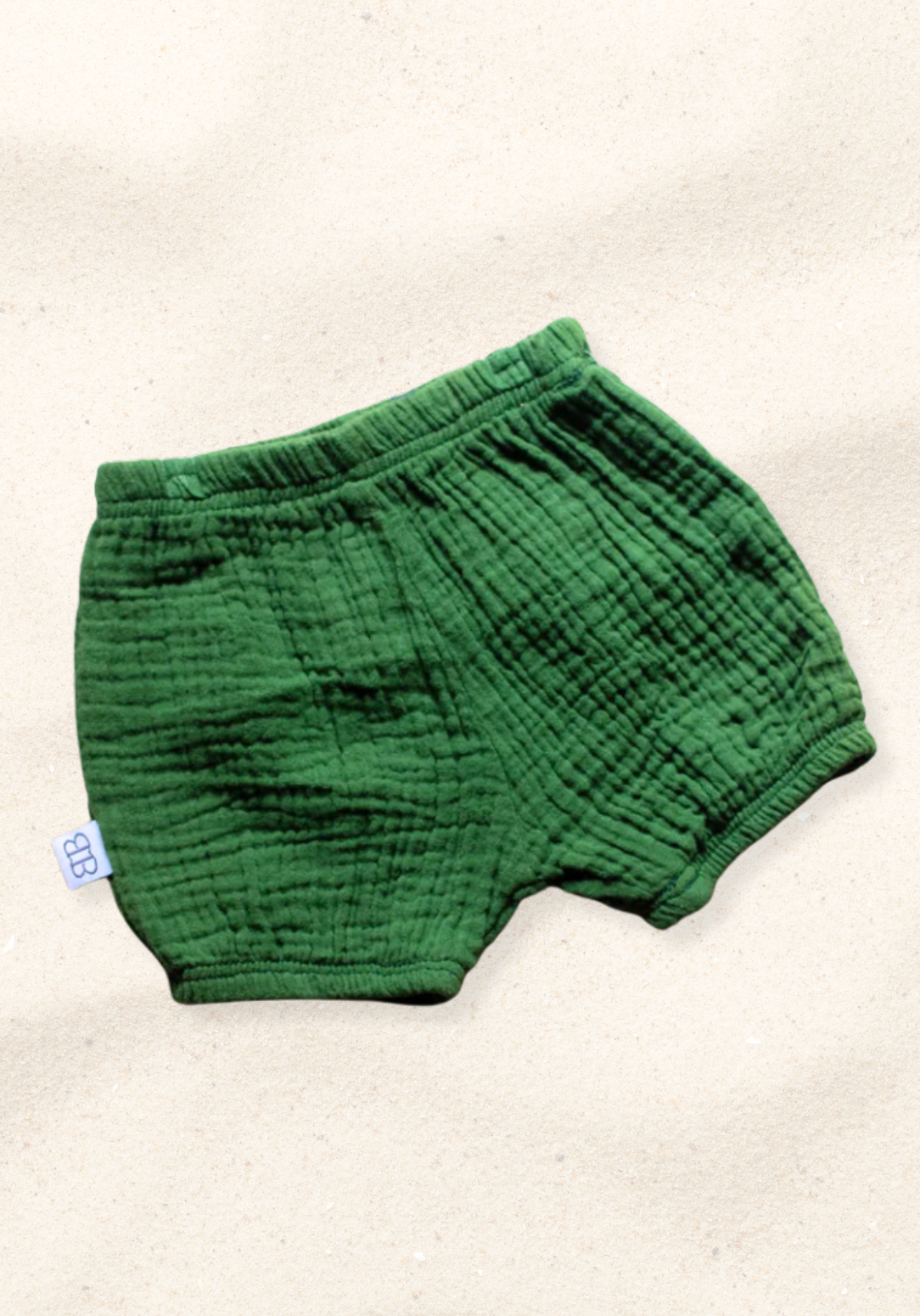 Deep green organic cotton baby bloomers with elastic waistband and leg openings, laid flat on a sand-colored background