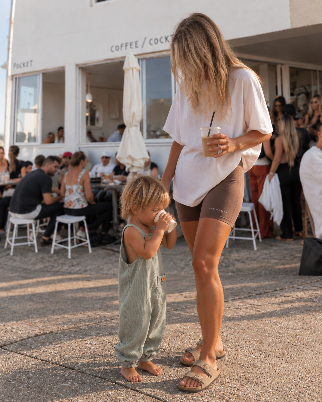 A woman and a young child standing outdoors at a cafe, with the woman wearing an oversized white t-shirt and fitted brown leggings, and the child wearing a light sage green sleeveless overalls jumpsuit.
